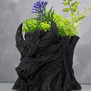 May include: A black, dragon-themed planter with a textured finish. The planter is filled with green and purple artificial plants. The dragon's head is prominently featured on the front of the planter, with horns and detailed scales.
