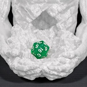 May include: A white, textured sculpture of a torso with outstretched arms cradling a green, multi-sided die. The die has white numbers and is used for tabletop games. The sculpture has a rough, organic appearance.