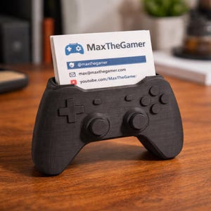 Può includere: Un porta biglietti da visita nero, stampato in 3D, a forma di controller per videogiochi. Il supporto contiene biglietti da visita con il testo "MaxTheGamer" e i profili dei social media. Il design del controller include pulsanti e un D-pad.