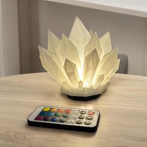 May include: A crystal-shaped lamp with a glowing interior, resting on a table. The lamp has a complex, faceted design, and a black base. A remote control with color options sits nearby.