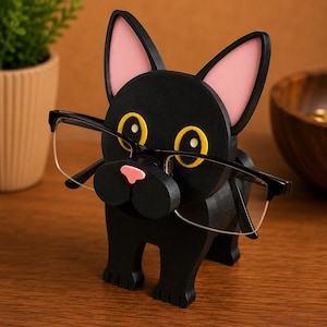 May include: A black wooden eyeglasses holder in the shape of a bulldog. The bulldog has pink inner ears, yellow eyes, and a pink nose. The holder is holding a pair of eyeglasses.