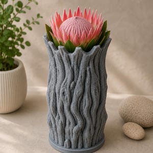 Op de afbeelding: Een grijze, getextureerde vaas in de vorm van een boomstam, met een roze protea bloem met groene bladeren. De vaas heeft een gedetailleerd, schorsachtig uiterlijk. Een kleine potplant en twee stenen staan op de achtergrond.