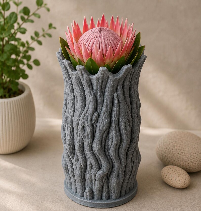 Op de afbeelding: Een grijze, getextureerde vaas in de vorm van een boomstam, met een roze protea bloem met groene bladeren. De vaas heeft een gedetailleerd, schorsachtig uiterlijk. Een kleine potplant en twee stenen staan op de achtergrond.