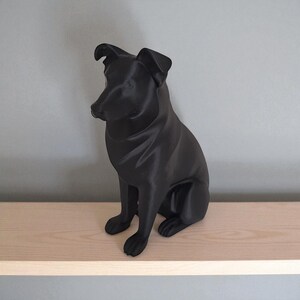 May include: A black dog sculpture in a seated position. The dog has a smooth, matte finish and is sitting on a light-colored wooden shelf. The sculpture has detailed features, including the ears, nose, and paws.
