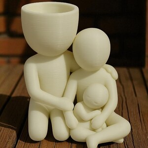 Puede incluir: Una figura familiar de color crema, que presenta una figura paterna con cabeza de maceta, abrazando a otro padre y a un ni&ntilde;o. Las figuras est&aacute;n estilizadas y parecen impresas en 3D, con un acabado liso y mate. La figura est&aacute; sobre una superficie de madera.