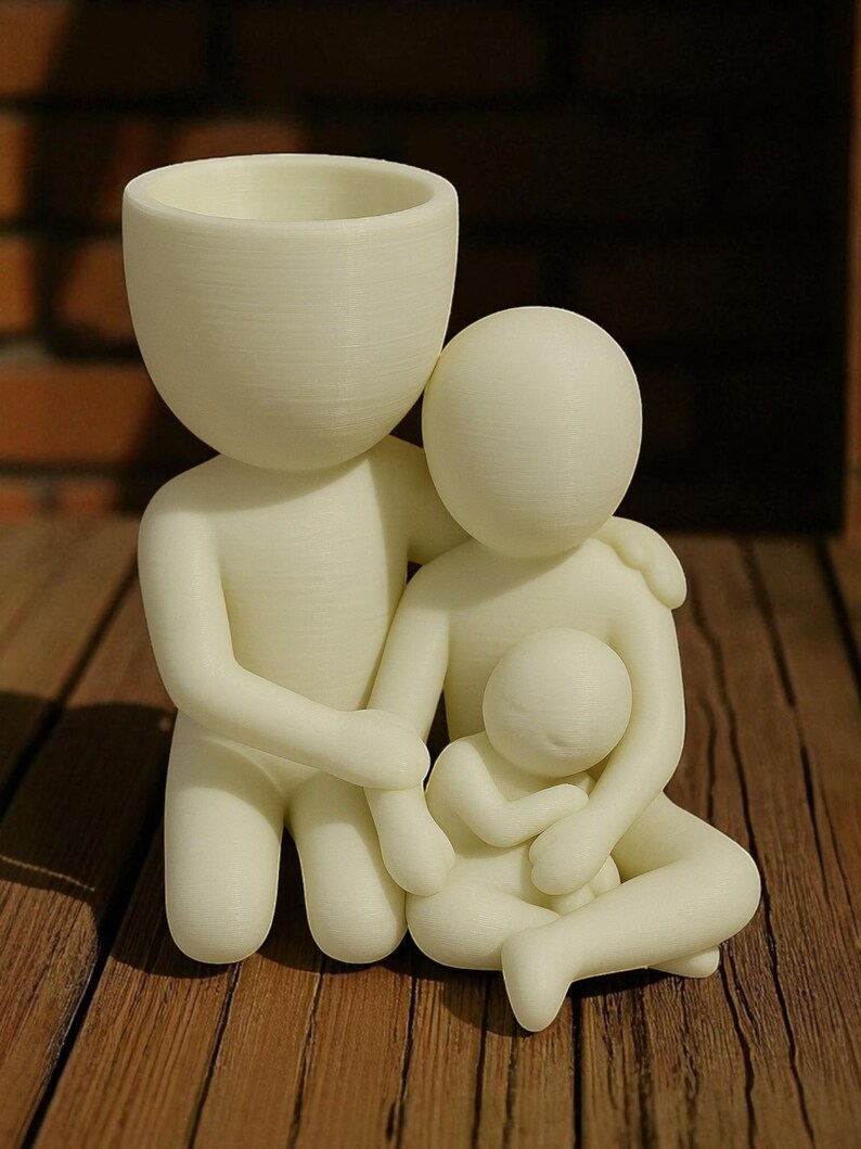 Puede incluir: Una figura familiar de color crema, que presenta una figura paterna con cabeza de maceta, abrazando a otro padre y a un ni&ntilde;o. Las figuras est&aacute;n estilizadas y parecen impresas en 3D, con un acabado liso y mate. La figura est&aacute; sobre una superficie de madera.