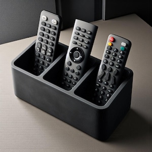 Puede incluir: Un organizador rectangular negro para mandos a distancia con tres compartimentos, que contiene tres mandos a distancia grises. Los mandos a distancia tienen numerosos botones. El organizador está sobre una superficie clara.