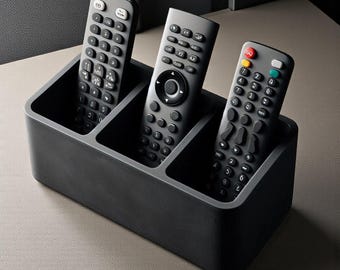 May include: A black rectangular remote control organizer with three compartments, holding three gray remote controls. The remotes have numerous buttons. The organizer sits on a light-colored surface.