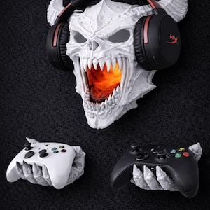 Può includere: Un supporto da parete a forma di teschio bianco con cuffie e bocca luminosa, che tiene due controller di gioco. I controller sono bianchi e neri. Le cuffie sono nere con dettagli rossi. Il teschio ha le corna.