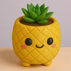 Puede incluir: Una maceta amarilla con forma de piña con una cara sonriente, ojos negros y mejillas rosadas. Contiene una planta suculenta verde. La maceta tiene un patrón de diamante texturizado y se apoya en tres pequeños pies.