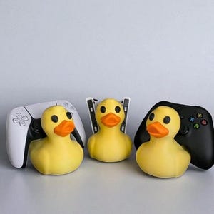 May include: Three yellow rubber duck toys with orange beaks and black eyes, each positioned with a different video game controller: a white PlayStation 5 controller, a PlayStation 5 console, and a black Xbox controller.