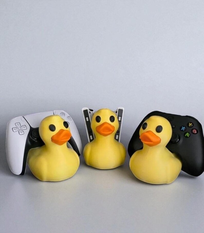 May include: Three yellow rubber duck toys with orange beaks and black eyes, each positioned with a different video game controller: a white PlayStation 5 controller, a PlayStation 5 console, and a black Xbox controller.