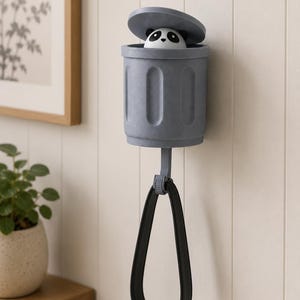 May include: A gray wall-mounted trash can-shaped holder with a panda bear figurine peeking out from the open lid. A black loop hangs below. The holder is mounted on a white paneled wall.