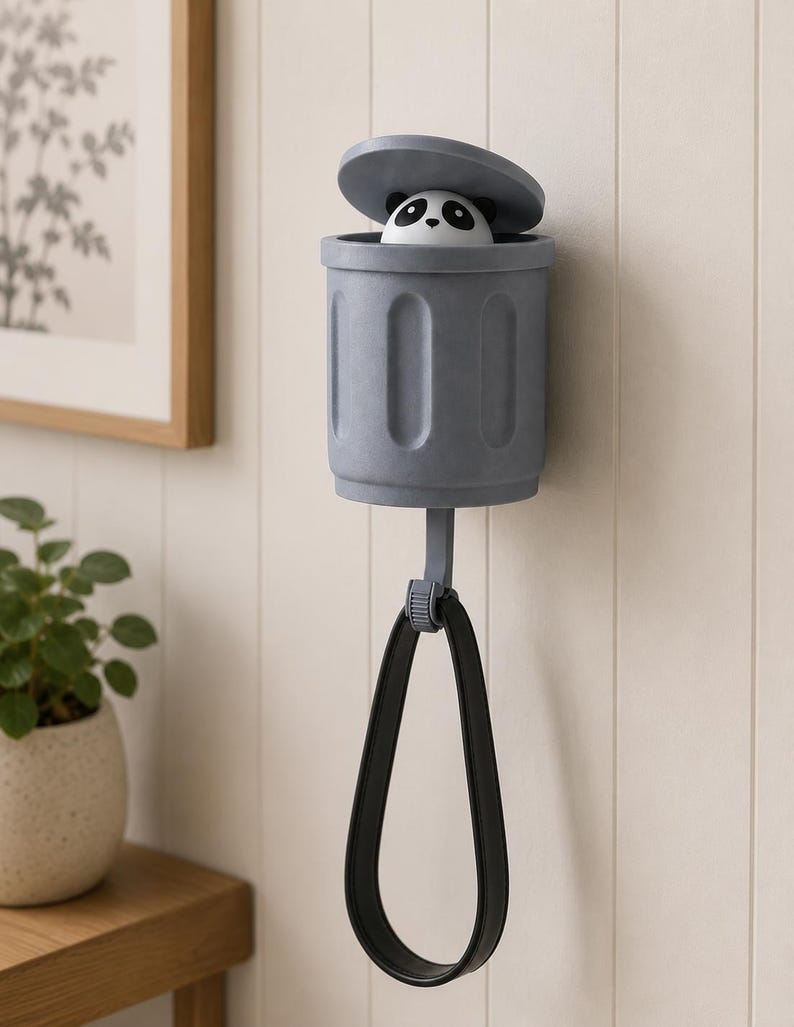 May include: A gray wall-mounted trash can-shaped holder with a panda bear figurine peeking out from the open lid. A black loop hangs below. The holder is mounted on a white paneled wall.