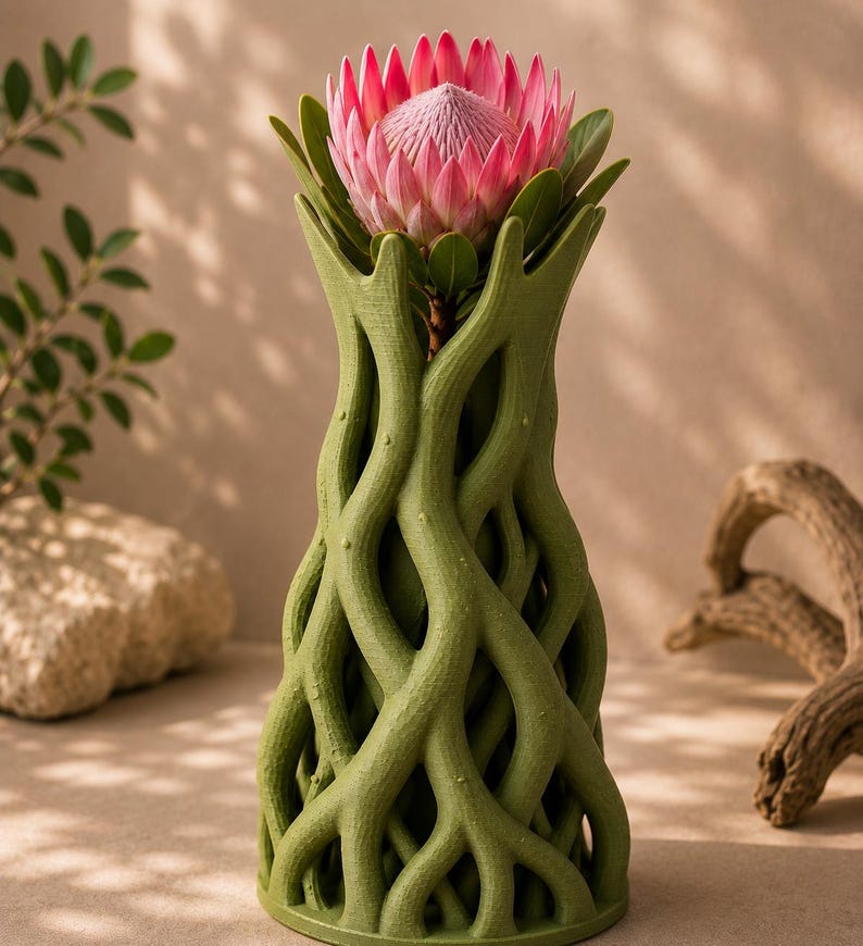 May include: A green, tree-root-inspired vase holding a pink protea flower with green leaves. The vase has a textured surface and a unique, organic design. The flower is in full bloom, showcasing its vibrant color.