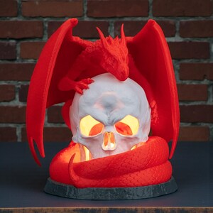 May include: A red dragon figurine perched atop a white skull with glowing yellow eyes and mouth. A red snake coils around the base. The dragon has outstretched wings. The sculpture sits on a dark gray base.