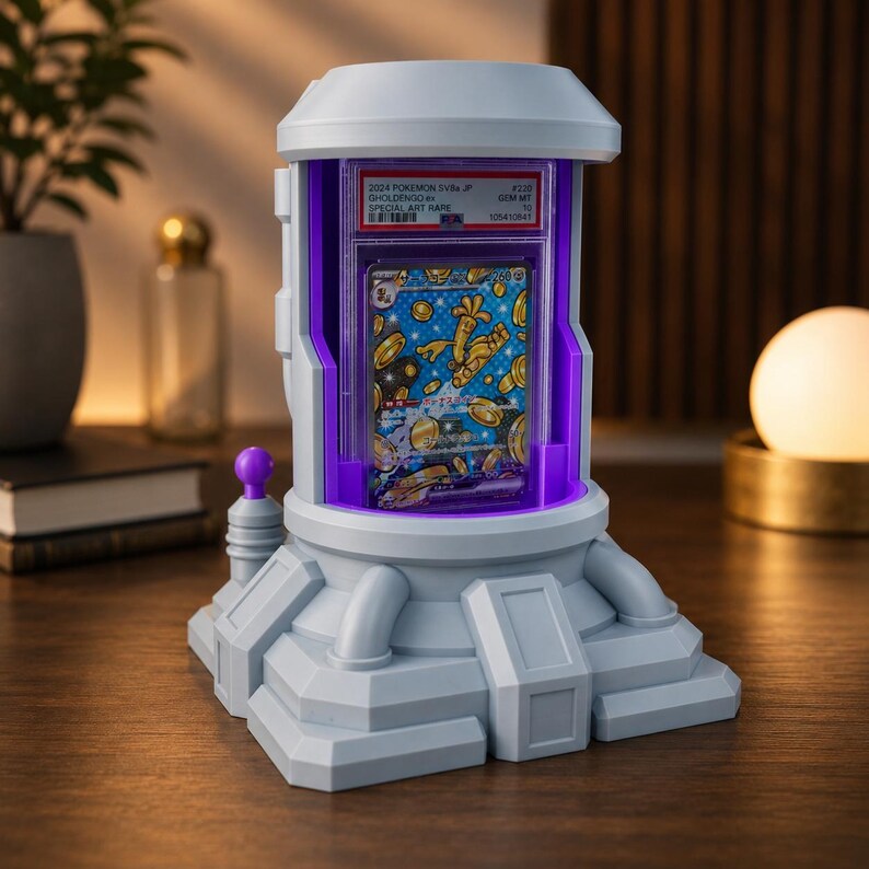 Op de afbeelding: Een grijze en witte display case met een paars interieur, met een Pok&eacute;mon trading card. De kaart heeft een blauwe achtergrond met gouden munten en een karakter. De case heeft een futuristisch ontwerp en is van binnenuit verlicht.