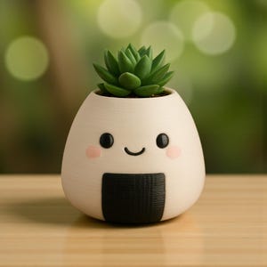 May include: A white, egg-shaped planter with a smiling face, black eyes, and pink cheeks, holding a green succulent plant. The planter has a black rectangular design at the bottom. The planter is sitting on a wooden surface.