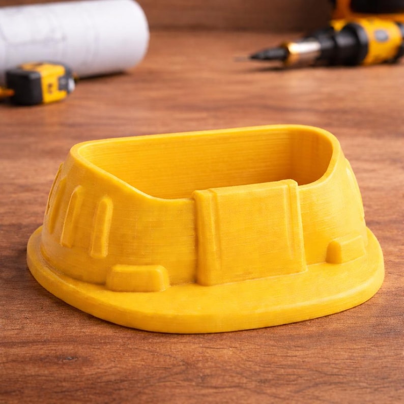May include: A bright yellow, 3D-printed container shaped like a construction worker's hard hat. The container has a rounded base and a rectangular opening. The hard hat design includes ridges and a front brim. The container sits on a wooden surface.