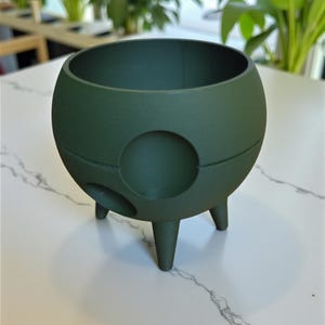 Puede incluir: Una maceta esf&eacute;rica verde oscuro con tres patas cortas y una abertura circular en el lateral. La maceta tiene un acabado liso y mate y una amplia abertura en la parte superior. El fondo presenta plantas verdes.
