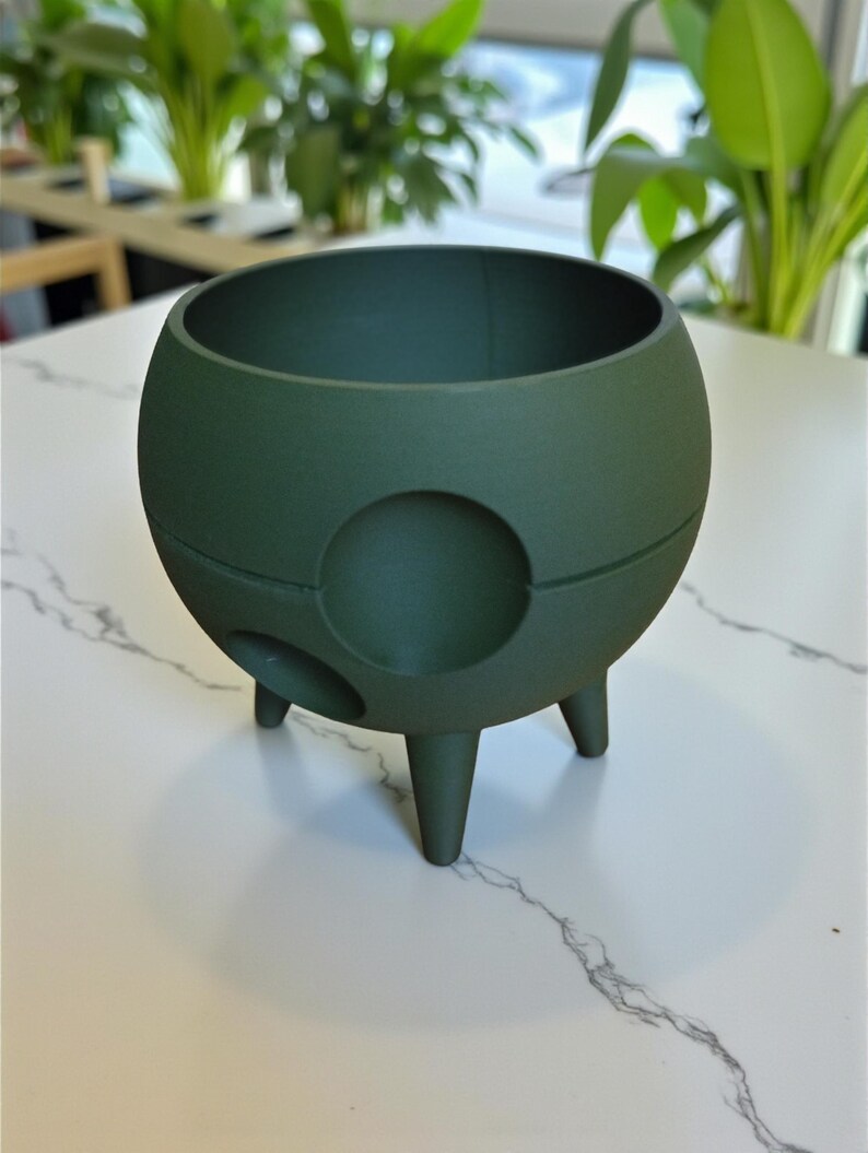 Puede incluir: Una maceta esf&eacute;rica verde oscuro con tres patas cortas y una abertura circular en el lateral. La maceta tiene un acabado liso y mate y una amplia abertura en la parte superior. El fondo presenta plantas verdes.