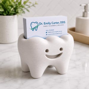Może przedstawiać: Biały, w kształcie zęba wizytownik z uśmiechniętą twarzą. Uchwyt zawiera stos wizyt&oacute;wek z tekstem "Dr. Emily Carter, DDS" i danymi kontaktowymi. Uchwyt znajduje się na białej powierzchni.