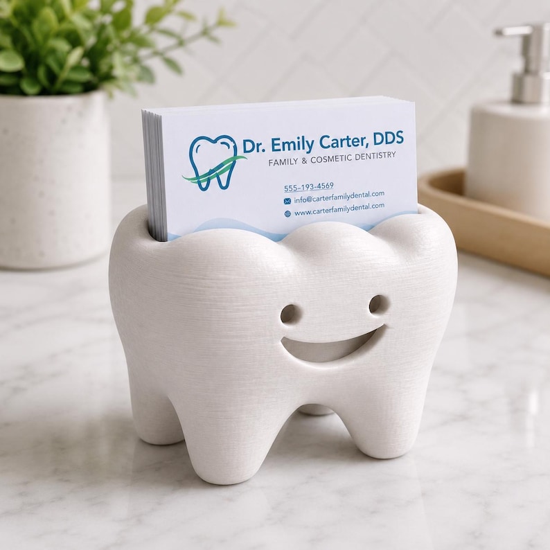 Może przedstawiać: Biały, w kształcie zęba wizytownik z uśmiechniętą twarzą. Uchwyt zawiera stos wizyt&oacute;wek z tekstem "Dr. Emily Carter, DDS" i danymi kontaktowymi. Uchwyt znajduje się na białej powierzchni.