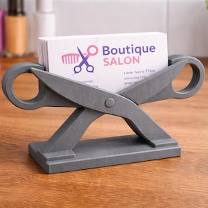 May include: A gray, scissors-shaped business card holder. The holder supports a stack of white business cards with the text "Boutique SALON" and a logo. The holder is made of a solid material and sits on a wooden surface.
