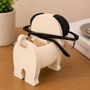 May include: A cream-colored pug-shaped glasses holder with black accents on the ears. Black-framed glasses rest on the holder, and a key ring hangs from the tail. The holder sits on a wooden surface, with a small plant and a book in the background.