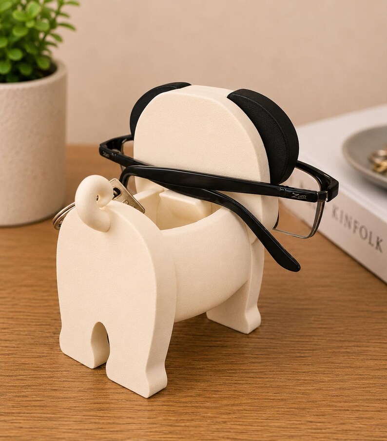 May include: A cream-colored pug-shaped glasses holder with black accents on the ears. Black-framed glasses rest on the holder, and a key ring hangs from the tail. The holder sits on a wooden surface, with a small plant and a book in the background.
