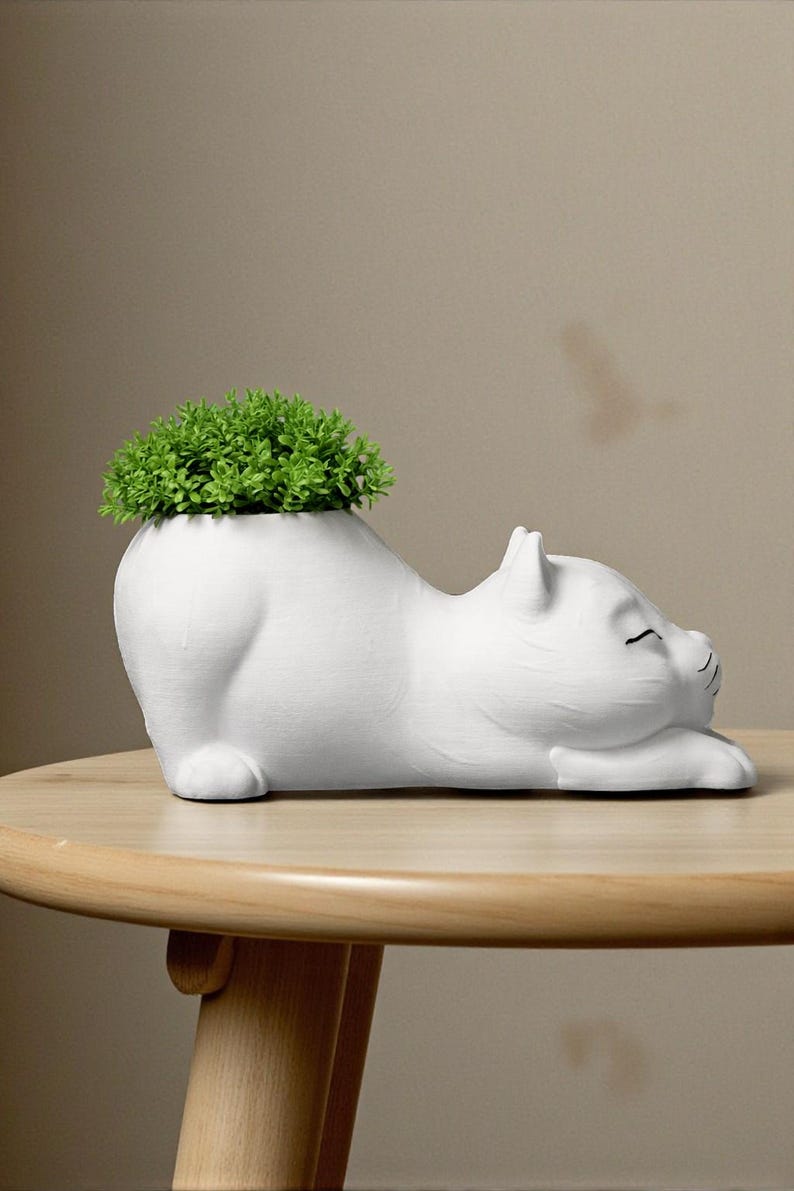 May include: A white ceramic cat-shaped planter with a green plant sprouting from its back. The cat is lying down with its eyes closed, and the planter sits on a light brown wooden table.