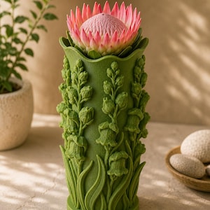 May include: A green vase with a detailed floral design, holding a pink protea flower. The vase has a textured surface with sculpted lily of the valley and leaf accents. The flower is a vibrant pink with a textured center.