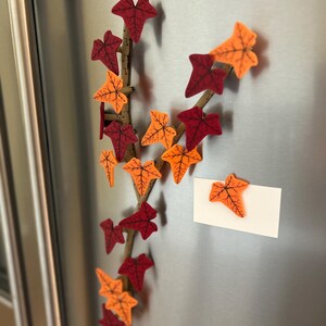 May include: A decorative fall-themed vine with orange and red leaves is attached to a silver refrigerator. A white note is attached to the refrigerator with a single orange leaf.
