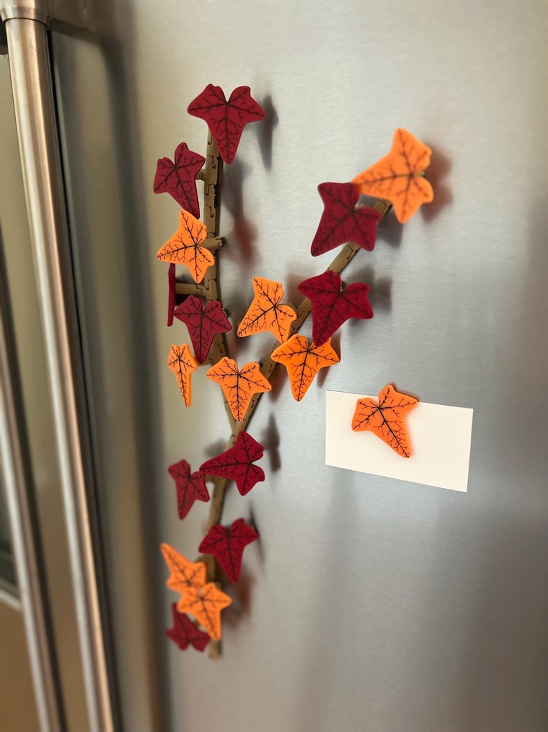 May include: A decorative fall-themed vine with orange and red leaves is attached to a silver refrigerator. A white note is attached to the refrigerator with a single orange leaf.