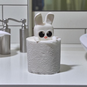 May include: A white bunny toilet roll holder with large black eyes and a pink nose sits on a roll of white, floral-patterned toilet paper. A decorative bathroom accessory.
