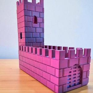 May include: A pink and purple toy castle with a tall tower and crenelated walls. The castle has a rectangular base and a small arched doorway with a gate. The tower has small windows and a similar crenelated top.