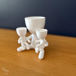 May include: A white ceramic planter with a large central pot and two smaller figures attached. The figures appear to be holding the smaller pots. The planter is on a wooden surface with a dark blue background.