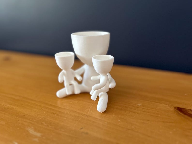 May include: A white ceramic planter with a large central pot and two smaller figures attached. The figures appear to be holding the smaller pots. The planter is on a wooden surface with a dark blue background.