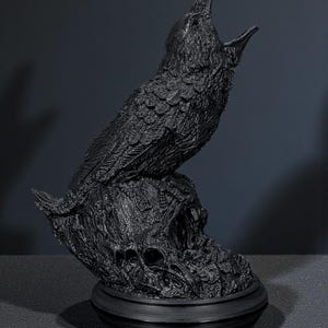 May include: A black raven sculpture with an open beak, perched atop a textured base with skull details. The raven is sculpted with detailed feathers. The sculpture is made of a dark material and sits on a round base.
