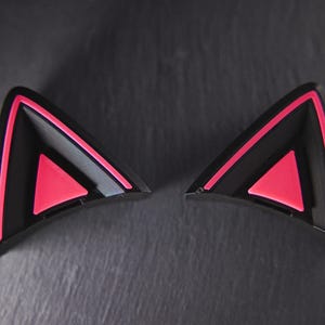 May include: A pair of black cat ears with pink accents. The ears are made of hard plastic and have a triangular shape.