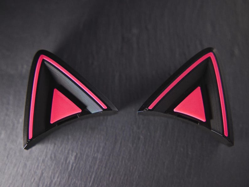 May include: A pair of black cat ears with pink accents. The ears are made of hard plastic and have a triangular shape.