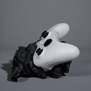 May include: A black and white controller stand shaped like a creature, holding a white Xbox controller. The stand has a textured surface and is designed to hold the controller in place. The background is a solid grey.