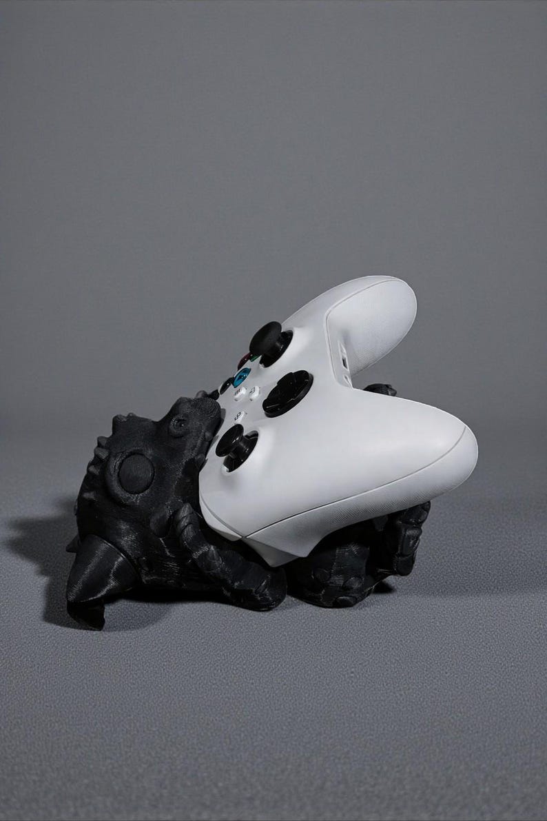 May include: A black and white controller stand shaped like a creature, holding a white Xbox controller. The stand has a textured surface and is designed to hold the controller in place. The background is a solid grey.