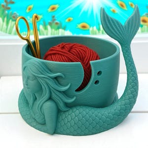 May include: Teal mermaid-themed yarn bowl with a red yarn ball inside. The bowl has a mermaid tail and a side profile of a mermaid. Gold crochet hooks and scissors are inside the bowl.
