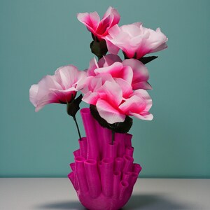 May include: A vibrant pink vase with a unique, coral-like design holds several pink and white artificial flowers. The flowers have dark green stems and leaves, and the background is a solid teal color. The vase is the focal point, showcasing a modern, artistic floral arrangement.