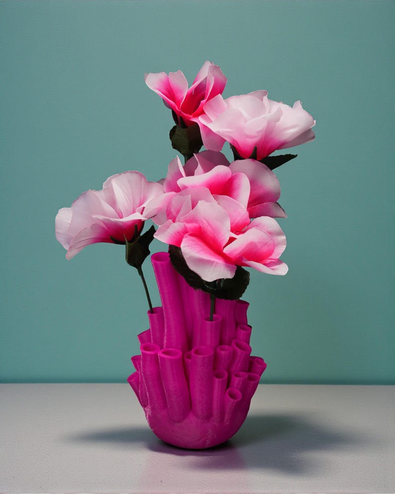 May include: A vibrant pink vase with a unique, coral-like design holds several pink and white artificial flowers. The flowers have dark green stems and leaves, and the background is a solid teal color. The vase is the focal point, showcasing a modern, artistic floral arrangement.