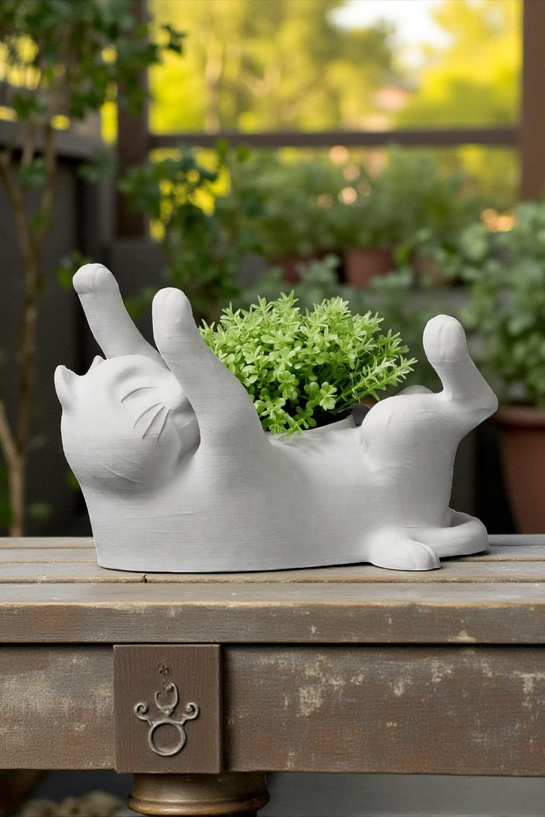 May include: A light gray cat-shaped planter, lying on its back with paws raised. The planter holds a small green plant. The cat is on a wooden surface, with a blurred background of greenery.