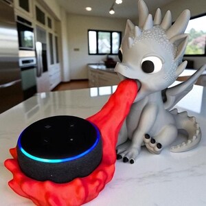 May include: A gray dragon-shaped speaker holder with a red flame-like design holding a black smart speaker. The speaker has a blue light ring. The dragon is sitting on a white surface.