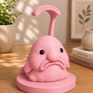 May include: A pink blobfish-shaped desk lamp with a curved handle. The lamp has black eyes and a sad expression. It sits on a round base. The lamp is made of a smooth material and is a unique decorative item.