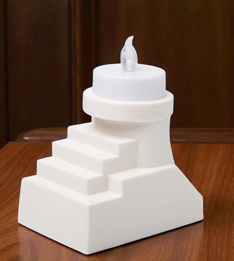 May include: A white, battery-operated LED candle with a realistic flickering flame. The candle is designed with a stepped base and a cylindrical top. The candle is made of a solid white material and is suitable for home decor.